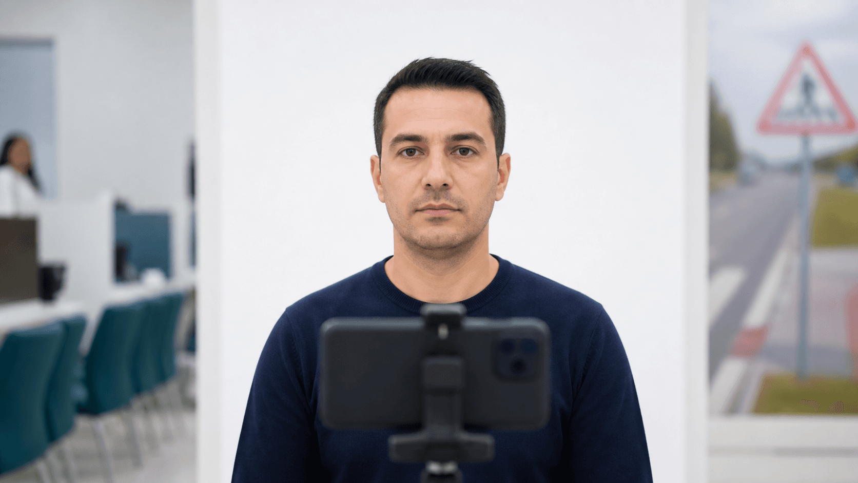 Sürücü belgesi başvurusu için beyaz fonda göz hizasına sabitlenmiş telefonla nötr ifadeli biyometrik fotoğraf çeken yetişkin